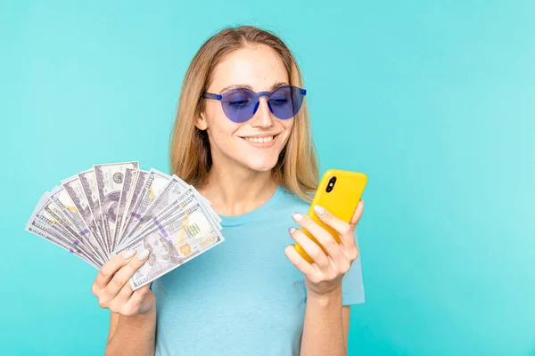 Jovem senhora isolado sobre fundo azul. Olhando câmera mostrando exibição de telefone celular segurando dinheiro . — Fotografia de Stock