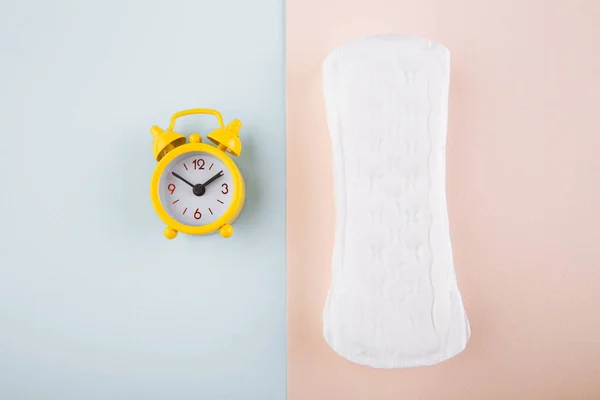 月経女性衛生の概念。ミニマルフラットレイアウト月経パッドと黄色の目覚まし時計上の青ピンクの背景 — ストック写真