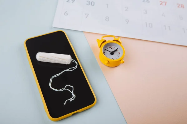 Smartphone to track your menstrual cycle and for marks. PMS and the critical days concept. Cotton tampon, daily pad and yellow alarm on the pink blue background