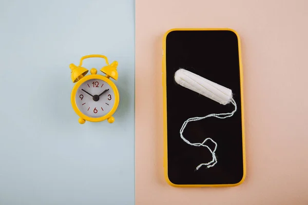 月经周期概念。带棉条的黄色闹钟和智能手机屏幕 — 图库照片