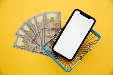 İnternetten alışveriş konsepti. Para, sepet ve akıllı telefon sarı arka planda izole edilmiş.