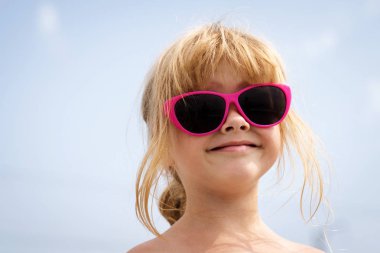 Gökyüzünde pembe güneş gözlüklü, gülümseyen küçük bir kızın büyük portresi.