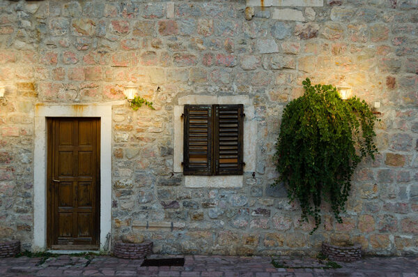 Facade of old house with a wooden door and a window, lit by two lanterns.Growing plants with foliage.