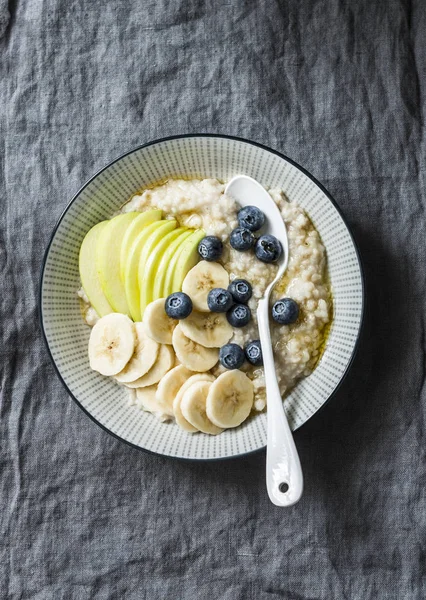 Steel-cut oats breakfast porridge with apple, banana, blueberry and honey on a grey background, top view