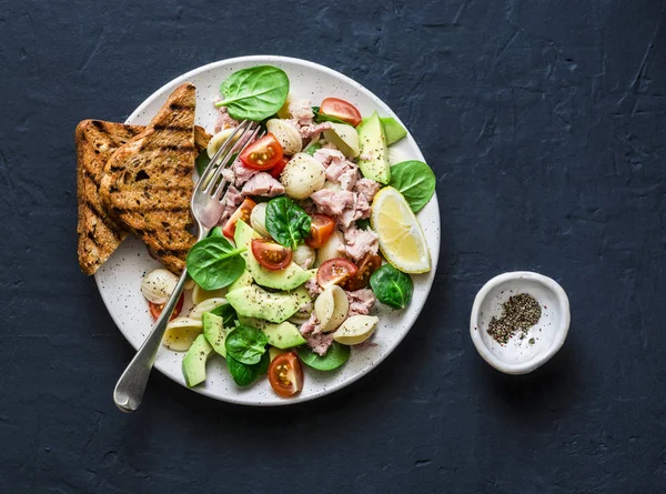 Orecchiette pasta, tuna, avocado, spinach, tomato salad and whole grain bread toast - delicious healthy lunch on a dark background, top view