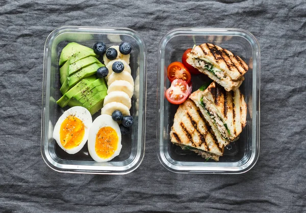 Two healthy office lunch box with sweet and savoury food. Boiled egg, avocado, tuna spinach cheese sandwiches and fruit on grey background, top view