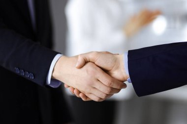 İş adamları toplantıda ya da pazarlıkta el sıkışıyor, yakın plan. Bir grup tanınmamış iş adamı ve modern bir ofiste duran bir kadın. Takım çalışması, ortaklık ve el sıkışma kavramı