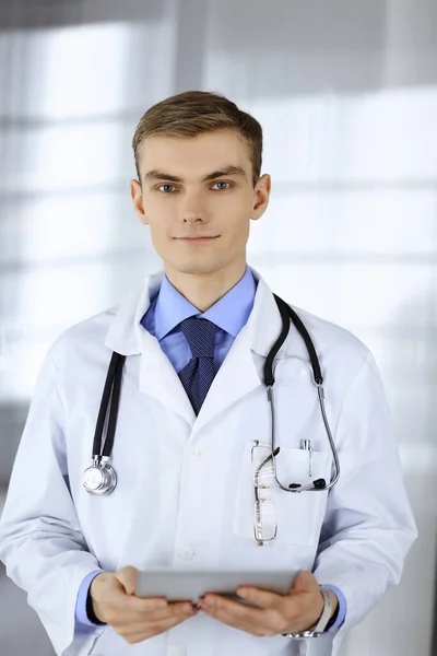 El joven doctor sonríe alegremente a la cámara, mientras está de pie en una oficina del hospital, sosteniendo una tableta de computadora en sus manos. Servicio médico perfecto en la clínica. Feliz futuro en la medicina y — Foto de Stock