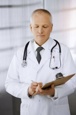 Yaşlı, kendine güvenen bir doktor güneşli klinikte dikilip pano kullanıyor. Mükemmel bir tıbbi servis. Tıp konsepti