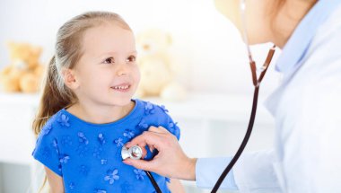 Güneşli klinikte bir çocuğu steteskopla muayene eden bir doktor. Mavi elbiseli mutlu gülümseyen kız hasta her zamanki sağlık kontrolünde.