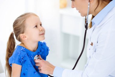 Güneşli klinikte bir çocuğu steteskopla muayene eden bir doktor. Mavi elbiseli mutlu gülümseyen kız hasta her zamanki sağlık kontrolünde.
