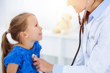 Güneşli klinikte bir çocuğu steteskopla muayene eden bir doktor. Mavi elbiseli mutlu gülümseyen kız hasta her zamanki sağlık kontrolünde.