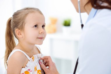 Güneşli klinikte bir çocuğu steteskopla muayene eden bir doktor. Parlak renkli elbise giymiş mutlu gülümseyen kız hasta her zamanki sağlık kontrolünde.