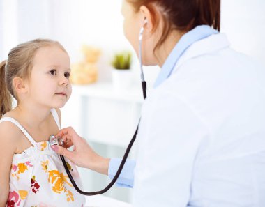 Güneşli klinikte bir çocuğu steteskopla muayene eden bir doktor. Parlak renkli elbise giymiş mutlu gülümseyen kız hasta her zamanki sağlık kontrolünde.