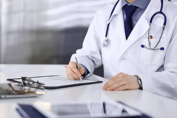 Unknown male doctor sitting and working with clipboard of medication history record in clinic at his working place, close-up. Young physician at work. Perfect medical service, medicine concept — Stock Photo, Image