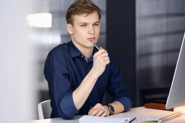 Jeune homme d'affaires blond réfléchissant à la stratégie à son lieu de travail avec ordinateur. Démarrage d'entreprise signifie travailler dur et hors du temps pour la réussite — Photo