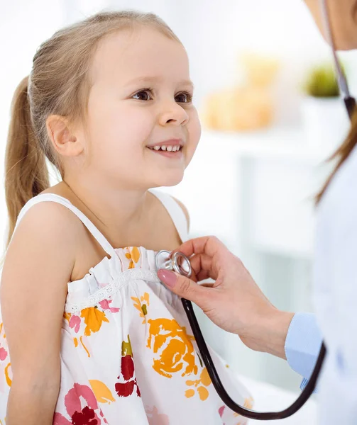 Doutor examinando uma criança por estetoscópio na clínica ensolarada. Feliz sorrindo paciente menina vestida com vestido de cor brilhante é na inspeção médica habitual — Fotografia de Stock