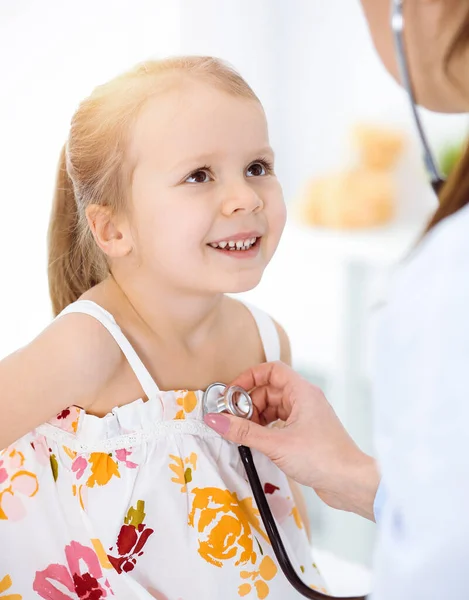 Doutor examinando uma criança por estetoscópio na clínica ensolarada. Feliz sorrindo paciente menina vestida com vestido de cor brilhante é na inspeção médica habitual — Fotografia de Stock