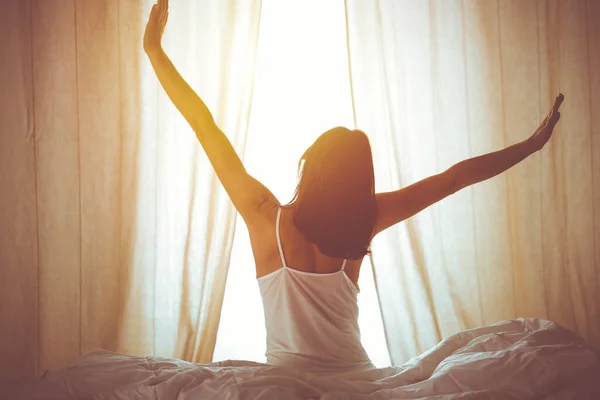 Mujer estirando las manos en la cama después de despertar, destello del sol. Morena entrando en un día feliz y relajado después de una buena noche de sueño y vista trasera. Concepto de un nuevo día y fin de semana alegre —  Fotos de Stock