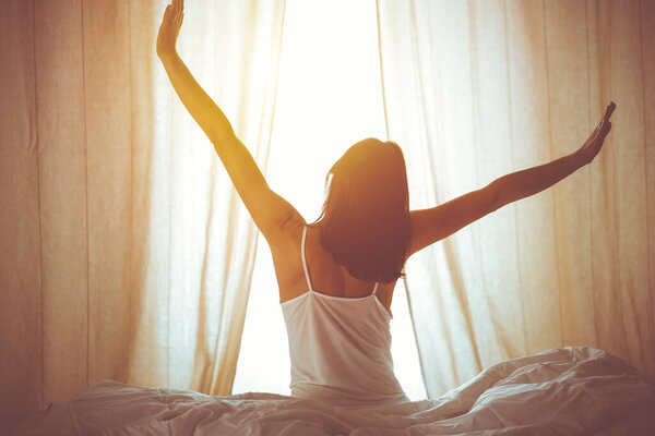 Woman stretching hands in bed after wake up, sun flare . Brunette entering a day happy and relaxed after good night sleep and back view. Concept of a new day and joyful weekend