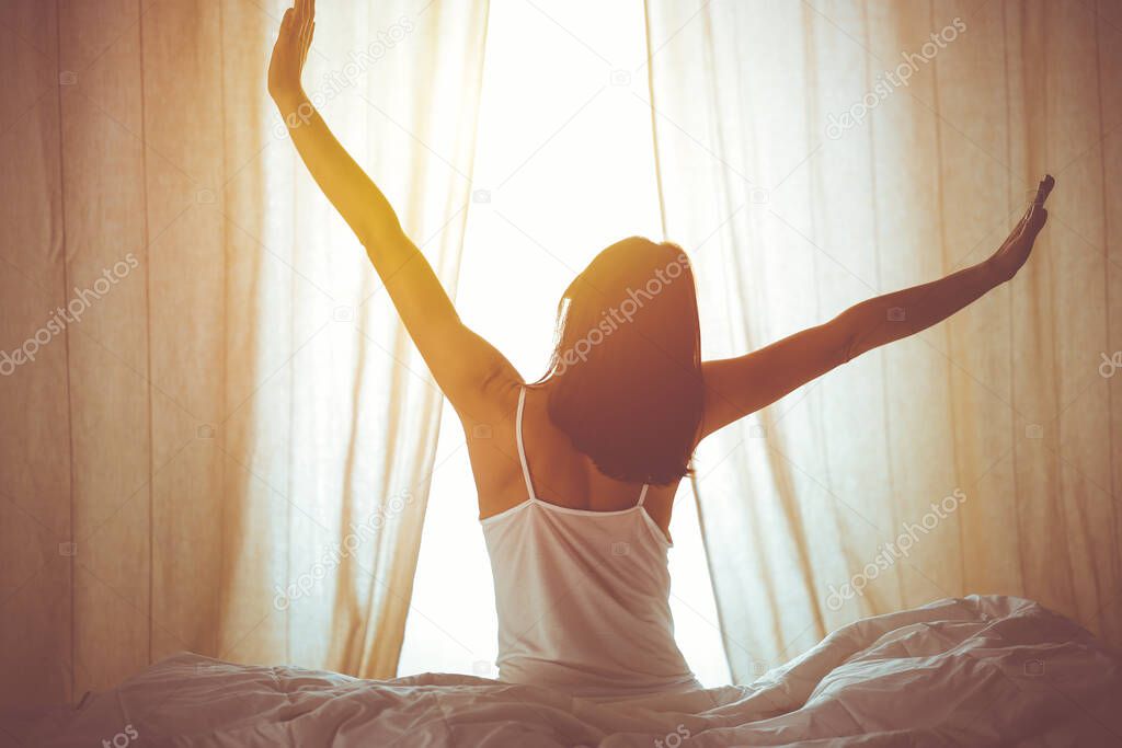 Woman stretching hands in bed after wake up, sun flare . Brunette entering a day happy and relaxed after good night sleep and back view. Concept of a new day and joyful weekend