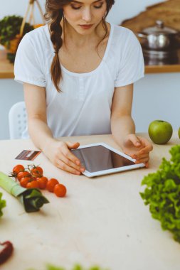 Genç bir kadın mutfakta yemek pişirmek için yeni bir tarif arıyor. Ev hanımı tablet bilgisayar ve kredi kartıyla online alışveriş yapıyor.
