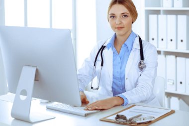 Hastanedeki genç bayan doktor masaüstü bilgisayar monitörüne bakıyor. Doktor ilaç geçmişi ve sınav sonuçlarını kontrol ediyor. Tıp ve sağlık hizmetleri kavramı