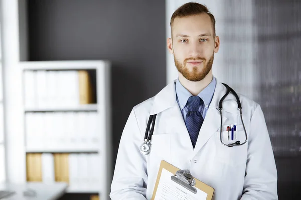 Medico con la barba rossa in piedi dritto in clinica vicino al suo posto di lavoro. Ritratto di medico. Medicina e concetto di assistenza sanitaria — Foto Stock