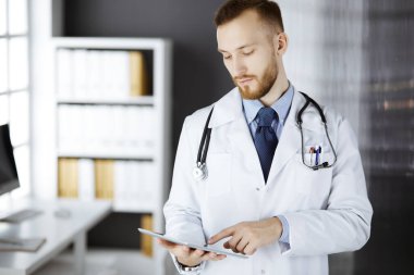 Dost canlısı kızıl sakallı doktor çalıştığı yerdeki klinikte tablet bilgisayar kullanıyor. Hastane ve tıp konseptinde mükemmel bir tıbbi hizmet.