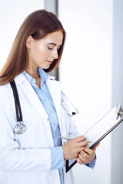 Ärztin füllt Arztformular aus, während sie am Fenster in der Klinik steht. Arzt bei der Arbeit. Medizin und Gesundheitskonzept — Stockfoto