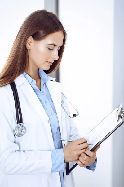 Medico donna riempiendo la forma medica mentre in piedi vicino alla finestra in clinica. Medico al lavoro. Medicina e concetto di assistenza sanitaria — Foto Stock