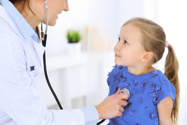 Doktor steteskopla küçük bir kızı muayene ediyor. Her zamanki tıbbi muayenede gülümseyen çocuk hasta. Tıp ve sağlık hizmetleri kavramları