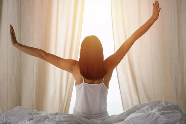 Kobieta wyciągająca ręce do łóżka po przebudzeniu, rozbłysk słońca. Brunetka wchodzi dzień szczęśliwy i zrelaksowany po dobrej nocy snu i widok z tyłu. Koncepcja nowego dnia i radosny weekend — Zdjęcie stockowe