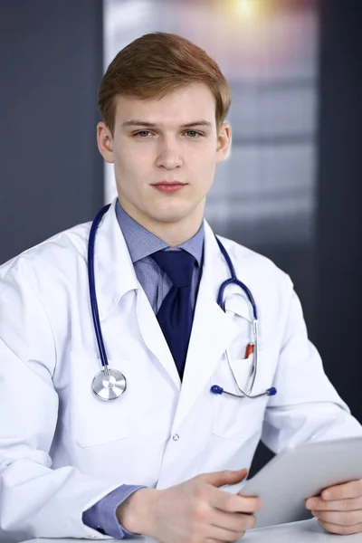 Blonder Arzt sitzt und arbeitet mit Tablet-Computer in sonniger Klinik an seinem Arbeitsplatz, in Großaufnahme. Medizinkonzept — Stockfoto