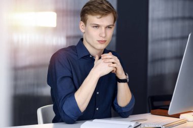 Genç sarışın işadamı, çalışma yerinde, karanlık bir ofiste bilgisayarla strateji düşünüyor. Arka planda ışık parıltısı var.