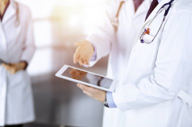 Bilinmeyen bir grup doktor, güneşli bir hastane ofisinde dururken bazı tıbbi isim kayıtlarını kontrol etmek için bir bilgisayar tableti kullanıyor. Doktorlar hastaları muayene etmeye ve yardım etmeye hazır. Tıbbi yardım, sigorta.