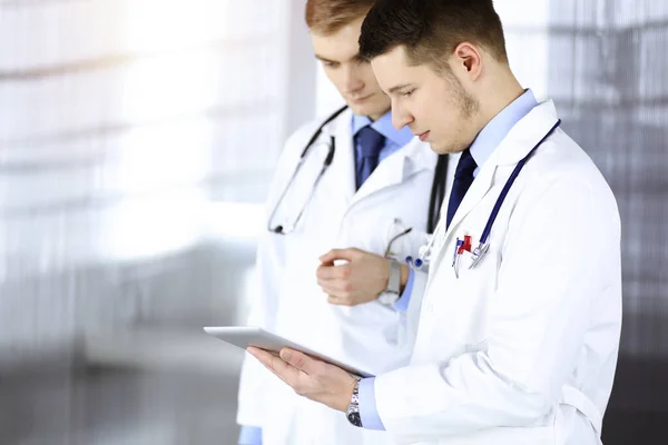 Twee artsen met stethoscopen bespreken medische onderzoeken die in het zonnige ziekenhuiskantoor staan. Artsen en zijn collega 's gebruiken een tablet voor het controleren van medicijnnamen. Perfecte medische — Stockfoto