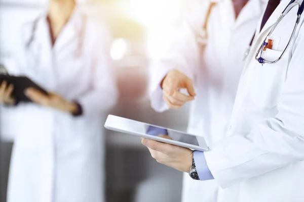 Group of unknown doctors use a computer tablet to check up some medical names records, while standing in a sunny hospital office. Physicians ready to examine and help patients. Medical help, insurance