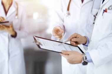Tanınmayan doktorlar, iki erkek ve bir kadın, güneşli hastane ofisinde dururken tıbbi muayene sonuçlarını tartışıyorlar. Doktorlar ilaç kayıtlarını doldurmak için pano kullanıyorlar. Mükemmel sağlık.