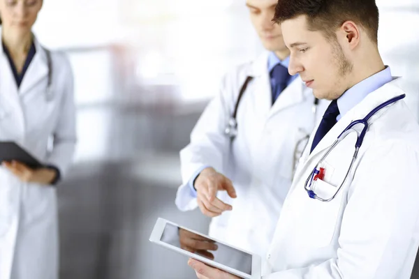 Ομάδα γιατρών ελέγχει τα ιατρικά ονόματα σε ένα tablet υπολογιστή, με μια νοσοκόμα με ένα πρόχειρο στο παρασκήνιο, στέκεται μαζί σε ένα ηλιόλουστο γραφείο νοσοκομείου. Γιατροί έτοιμοι να εξετάσουν και να βοηθήσουν — Φωτογραφία Αρχείου