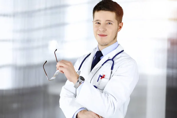 Dottore uomo con gli occhiali, è in piedi con un sorriso al suo soleggiato ufficio ospedaliero. Servizio medico perfetto in clinica. Buon futuro in medicina e sanità — Foto Stock