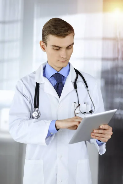 Young doctor man is standing in a sunny hospital office, holding a computer tablet in his hands. Perfect medical service in clinic. Happy future in medicine and healthcare