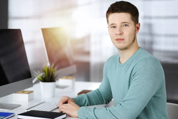 Der junge selbstbewusste Geschäftsmann sitzt in einem grünen Pullover am Schreibtisch in einem sonnigen modernen Büro, während er am Computer an seinem neuen Projekt arbeitet. Kopfschuss eines männlichen Unternehmers oder IT-Spezialisten — Stockfoto