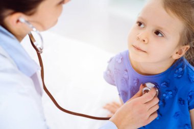 Güneşli klinikte bir çocuğu steteskopla muayene eden bir doktor. Mavi elbiseli mutlu gülümseyen kız hasta her zamanki sağlık kontrolünde.