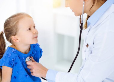 Güneşli klinikte bir çocuğu steteskopla muayene eden bir doktor. Mavi elbiseli mutlu gülümseyen kız hasta her zamanki sağlık kontrolünde.