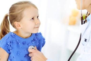 Güneşli klinikte bir çocuğu steteskopla muayene eden bir doktor. Mavi elbiseli mutlu gülümseyen kız hasta her zamanki sağlık kontrolünde.