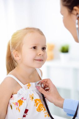 Güneşli klinikte bir çocuğu steteskopla muayene eden bir doktor. Parlak renkli elbise giymiş mutlu gülümseyen kız hasta her zamanki sağlık kontrolünde.