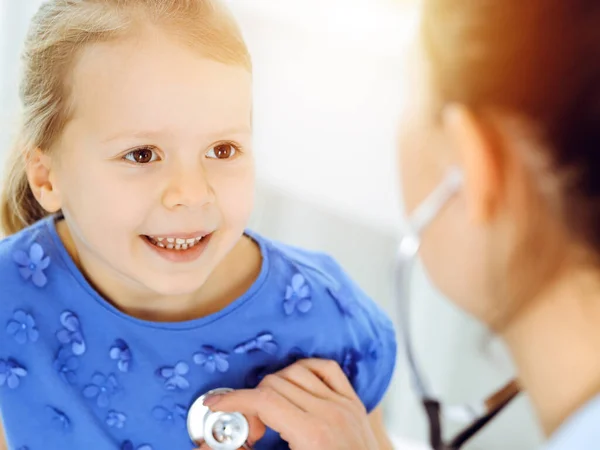 Doutor examinando uma criança por estetoscópio na clínica ensolarada. Feliz sorrindo paciente menina vestida de vestido azul é na inspeção médica habitual — Fotografia de Stock