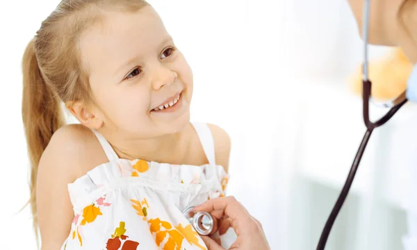 Врач осматривает ребенка стетоскопом в солнечной клинике. Счастливая улыбающаяся пациентка, одетая в яркое платье цвета при обычном медицинском осмотре — стоковое фото