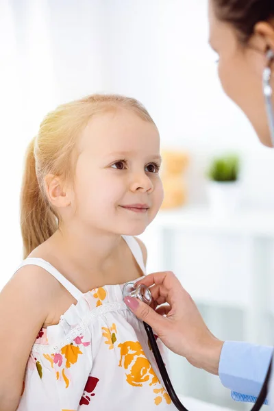 Doutor examinando uma criança por estetoscópio na clínica ensolarada. Feliz sorrindo paciente menina vestida com vestido de cor brilhante é na inspeção médica habitual — Fotografia de Stock